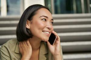 Cellular technology. Smiling asian girl talks on mobile phone, makes a call. Young woman with telephone sits on stairs photo