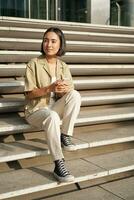 Beautiful asian girl sitting on stairs outside building, using mobile phone, looking at smartphone app and smiling photo