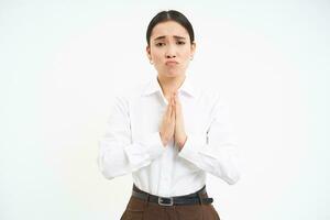 Sad and miserable asian woman, holds hands in pray, pleading, begging for help, asking you for favour, white background photo