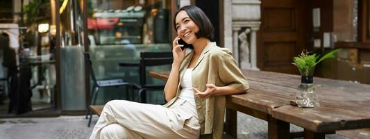 joven mujer teniendo conversacion en móvil teléfono, sentado al aire libre y haciendo teléfono llamar, utilizando teléfono inteligente, hablando foto