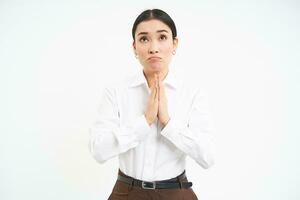 Hopeful corporate woman holds hand in pray, makes wish, looks up with hopeful face, standing over white background photo