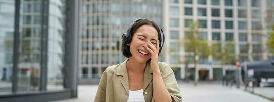 Carefree asian girl, laughing and smiling, wearing headphones and walking on street. Outdoor shot of young woman listening music and looking happy photo