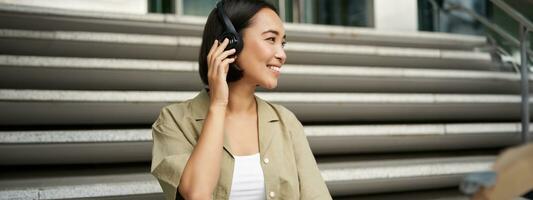 Close up portrait of smiling asian woman listens music in headphones, turns around with happy face expression, resting outdoors photo