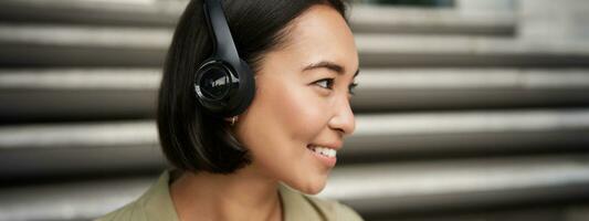 Close up portrait of smiling asian girl in headphones, listens to music outdoors, looking happy. photo