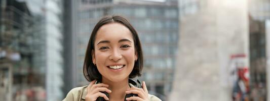 Vertical shot of beautiful asian woman posing with headphones around neck, smiling and laughing, standing on street in daylight photo