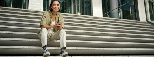 Portrait of smiling asian girl sits on stairs outdoors, sending message, using smartphone app, looking at mobile phone screen photo