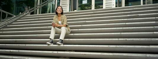 Portrait of smiling asian girl sits on stairs outdoors, sending message, using smartphone app, looking at mobile phone screen photo