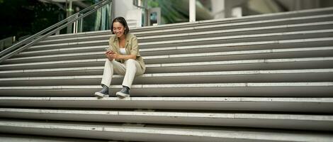 Portrait of smiling asian girl sits on stairs outdoors, sending message, using smartphone app, looking at mobile phone screen photo