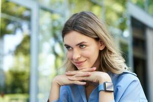 cerca arriba retrato de sonriente rubio mujer, estudiante con digital mirar, mirando contento a cámara, sentado fuera de en calle foto