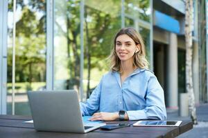 Portrait of young beautiful working woman, works on remote with laptop. Student takes notes during online video conference, sitting on street with computer photo