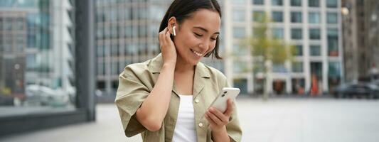 Smiling asian girl listens music in wireless headphones, looks at her phone, choosing music or podcast. Young woman calling someone, using headphones photo