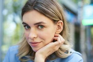 Close up portrait of young woman, tuck hair behind ear, looking flirty and smiling, sitting in blue shirt outdoors on street photo