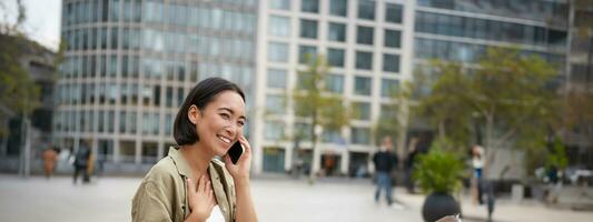 Vertical shot of smiling stylish asian girl, having a telephone conversation and walking on street. Young woman talking on mobile phone photo