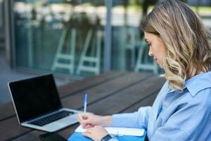 Close up portrait of woman student attend online course classes, sitting outside on fresh air with laptop and taking notes. Businesswoman video chat on computer photo