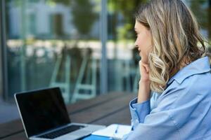 cerca arriba retrato de mujer estudiante atender en línea curso clases, sentado fuera de en Fresco aire con ordenador portátil y tomando notas mujer de negocios vídeo charla en computadora foto