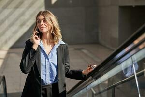 Smiling successful business woman talking on mobile phone while going up on escalator, walking in city photo