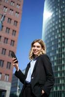 Vertical shot of successful businesswoman in suit, holding smartphone, using mobile phone on her way to work, standing with telephone on street photo