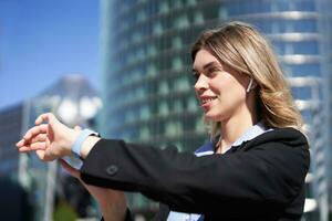 Portrait of busy corporate woman in wireless headphones, looking at time on her digital watch, checking messages, adjusting music, standing outdoors on street photo