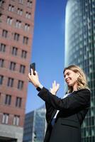 Portrait of smiling corporate woman video call on street, holding mobile phone and waving at smartphone camera, wearing suit photo