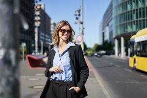 Portrait of confident corporate woman, young saleswoman in suit and sunglasses, looking self-assured on city street photo