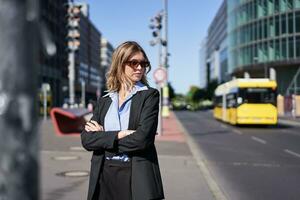 retrato de confidente negocio mujer en traje, cruzar brazos en pecho, mirando seguro de sí mismo en ciudad centro, en pie en calle foto