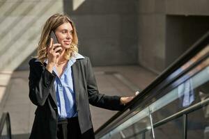 Smiling successful business woman talking on mobile phone while going up on escalator, walking in city photo