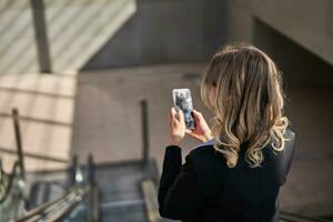 retrato de corporativo mujer en pie en escalera mecánica, haciendo un teléfono llamar, utilizando teléfono inteligente, caminando en ciudad foto