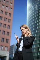 Portrait of successful businesswoman having a telephone conversation in wireless headphones, walking on street. Corporate woman with smartphone in city center photo