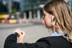 Portrait of busy corporate woman in wireless headphones, looking at time on her digital watch, checking messages, adjusting music, standing outdoors on street photo