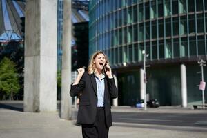 emocionado mujer de negocios recibe genial noticias, celebrando mientras hablando en móvil teléfono. corporativo mujer gritos desde alegría en calle, triunfando foto