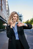 Portrait of confident businesswoman going on a meeting, standing on street, talking on mobile phone and checking time on digital watch photo