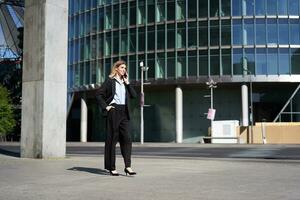 Young corporate woman in suit walking on street and talking on mobile phone. Businesswoman having telephone conversation while going to work photo