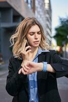 Vertical portrait of businesswoman having a phone call, looking at time on her digital watch, reading message on device photo