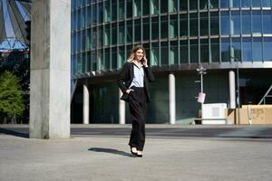 Young corporate woman in suit walking on street and talking on mobile phone. Businesswoman having telephone conversation while going to work photo