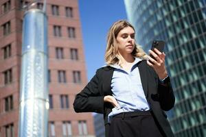 Low angle shot of businesswoman in suit, standing on street and looking at mobile phone, holding smartphone photo