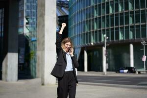 emocionado mujer de negocios recibe genial noticias, celebrando mientras hablando en móvil teléfono. corporativo mujer gritos desde alegría en calle, triunfando foto