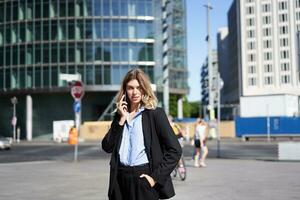 retrato de mujer de negocios en traje, en pie en calle con móvil teléfono, en llamada con alguien, sonriente mientras teniendo teléfono conversacion foto