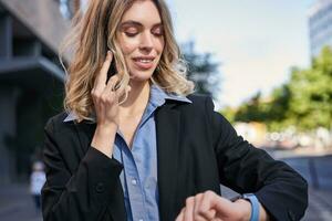 Portrait of confident businesswoman going on a meeting, standing on street, talking on mobile phone and checking time on digital watch photo