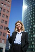 Vertical shot of successful businesswoman in suit, holding smartphone, using mobile phone on her way to work, standing with telephone on street photo