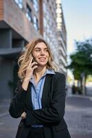 vertical Disparo de confidente joven mujer de negocios haciendo teléfono llamar, en pie en calle y hablando en teléfono foto