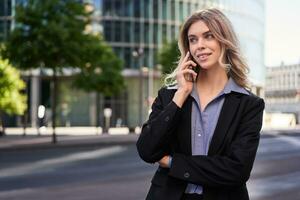 retrato de exitoso mujer de negocios, empresa CEO camina en calle y hace teléfono llamadas, hablando en teléfono inteligente foto