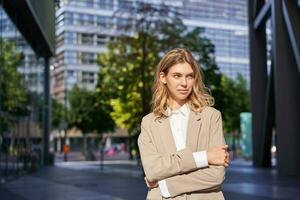 Portrait of young saleswoman, confident businesswoman in suit, cross arms on chest, standing in power pose on street near office buildings photo