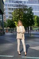 Portrait of young confident corporate woman in beige suit, standing on street in her business outfit, looking self-assured photo