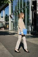 retrato de mujer de negocios en beige traje y alto tacones caminando en calle. corporativo mujer yendo a trabajar, participación carpeta con documentos foto