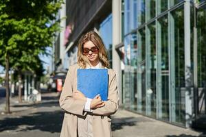 retrato de corporativo mujer en Gafas de sol y beige traje, participación azul carpeta con oficina documentos, yendo a trabajar, caminando en calle en soleado día foto