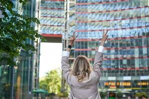 Corporate people. Rear view of businesswoman raising hands up in celebration, triumphing, achieve goal and rejoicing, standing on street photo