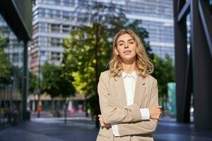 Portrait of young saleswoman, confident businesswoman in suit, cross arms on chest, standing in power pose on street near office buildings photo