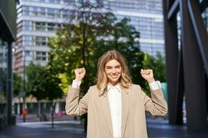 Portrait of successful business woman, young corporate woman celebrating victory, achievement, triumphing on street, making fist pumps and rejoicing photo