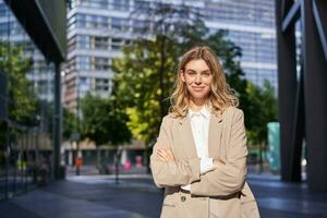 Portrait of young saleswoman, confident businesswoman in suit, cross arms on chest, standing in power pose on street near office buildings photo