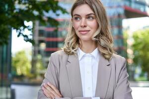 Portrait of young successful businesswoman in suit, cross arms on chest, smile and look confident, standing on street of business center photo